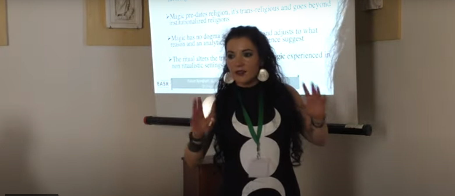 Dr Puca presenting in front of a screen wearing a black dress with a triple moon motif.